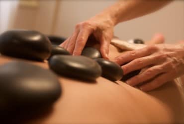 A soothing massage scene is presented, featuring smooth black stones placed on a person's back. Gentle hands are seen adjusting the stones, highlighting a serene atmosphere focused on relaxation and wellness.