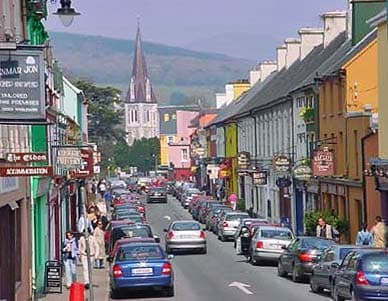 A vibrant street scene in Kenmare features a row of colorful buildings with shops and restaurants. Cars are parked along the road, and pedestrians are seen strolling. A church steeple rises in the background, framed by rolling hills against a clear sky.