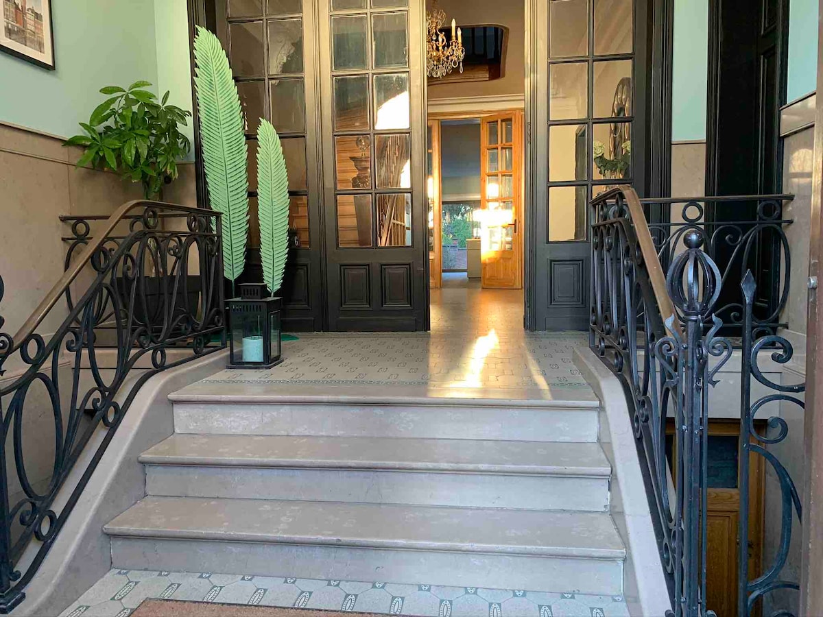 An elegant entrance is framed by ornate black railings and a potted plant. The staircase leads up to an inviting space illuminated by natural light, with a chandelier visible in the background. The tiled floor features a subtle pattern, enhancing the entry's classic charm.