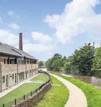 A stone building is set at canal level, featuring large windows that reflect natural light. A grassy area extends along the side, leading to a paved tow path bordered by lush greenery, providing a scenic route beside the tranquil water.