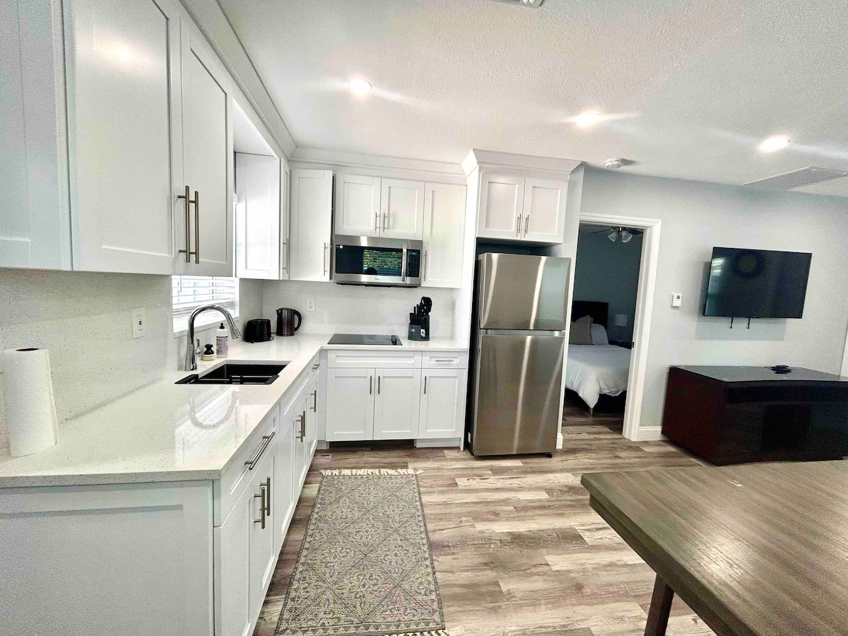 A modern kitchen is featured, showcasing sleek white cabinetry, stainless steel appliances, and a spacious countertop. Natural light brightens the space, while a dining table is positioned nearby. The open layout allows for easy access to the living area, visible in the background.
