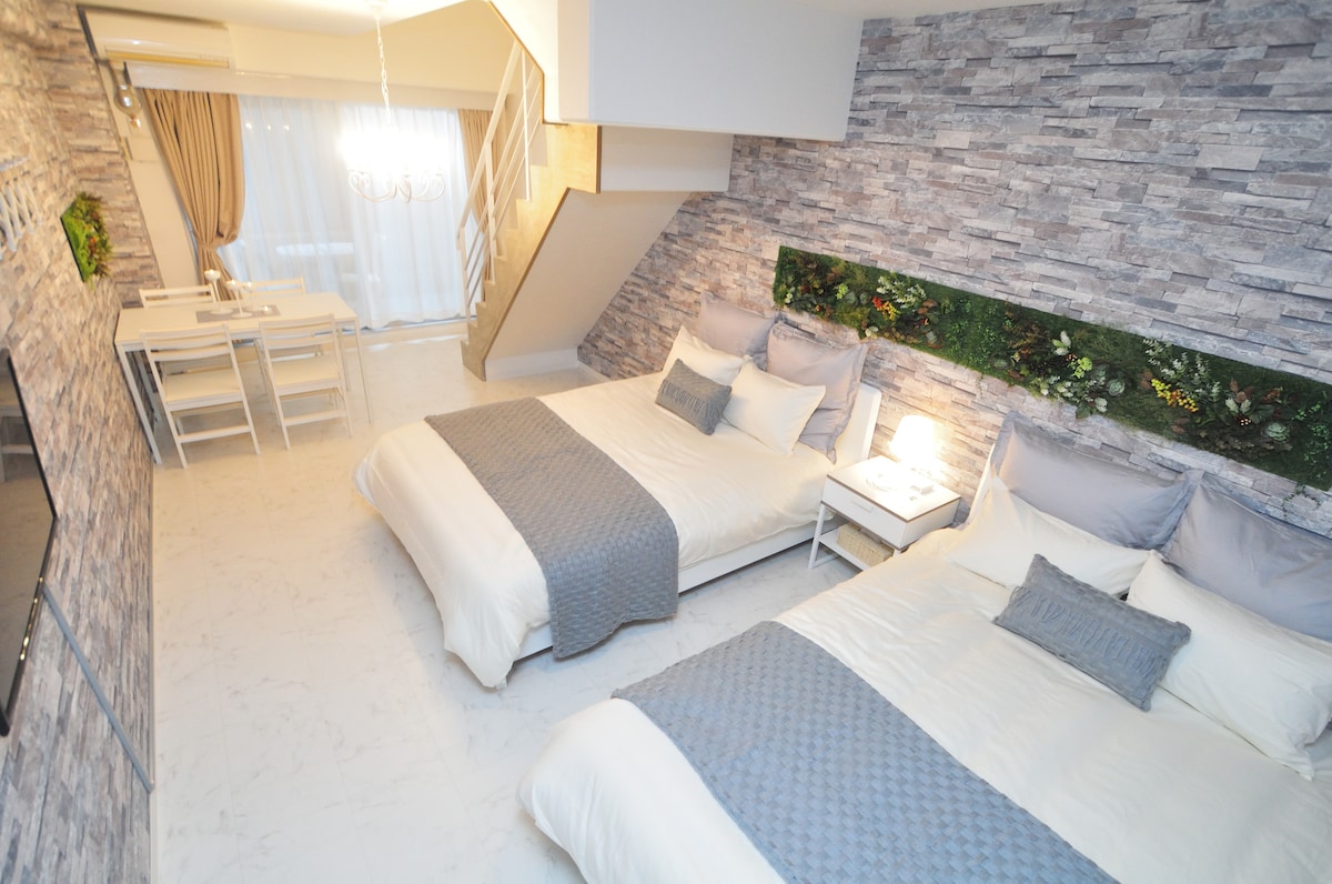 A spacious bedroom showcases two double beds with stylish gray bedding. A decorative stone wall adds texture, while a small bedside lamp provides soft illumination. In the background, a dining area can be seen, featuring a white table and chairs, along with a staircase leading to the upper level.