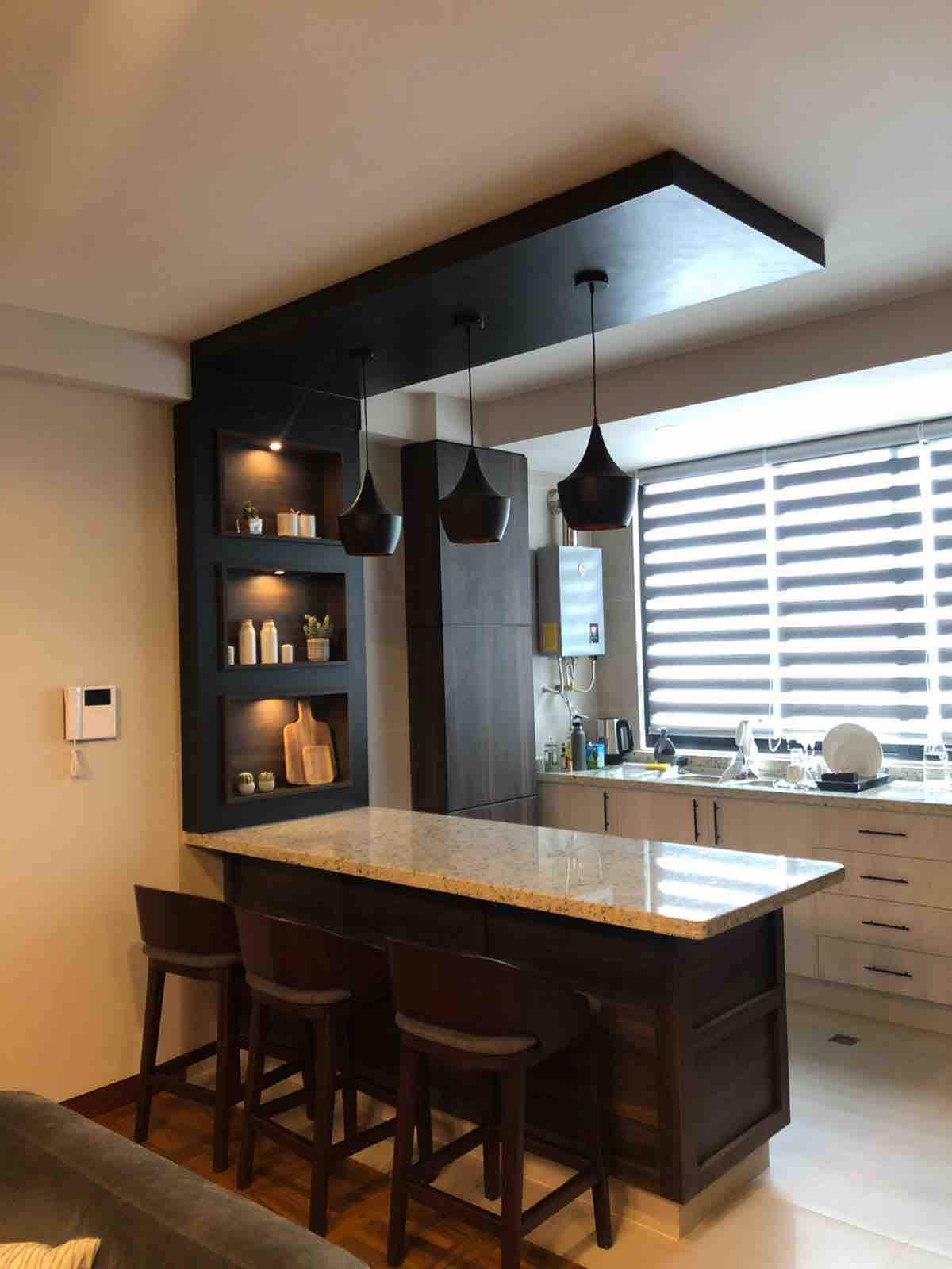 A modern kitchen is visible, featuring a granite countertop with three wooden stools positioned around it. Overhead, pendant lights accent a dark wood frame, while a window with striped blinds allows natural light. Shelves display various kitchen items, adding both function and style.