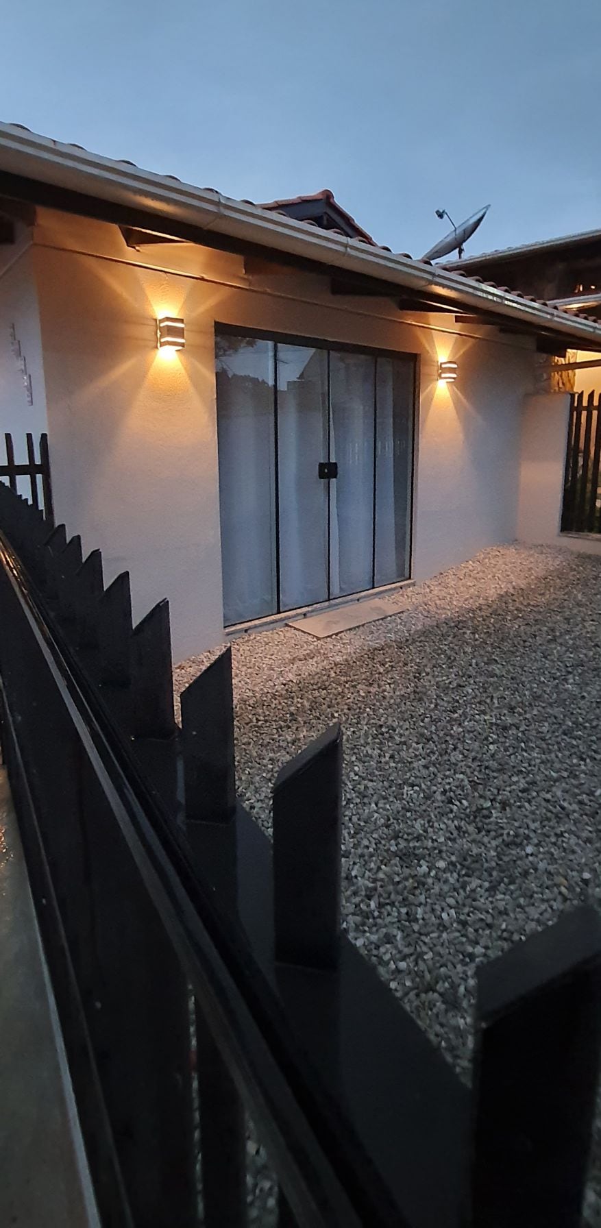 The exterior of the property features a gravel pathway leading to a glass door, framed by elegant lighting fixtures that enhance the entrance's visibility. A fence surrounds the area, offering privacy and security. The overall structure presents a clean and welcoming facade.