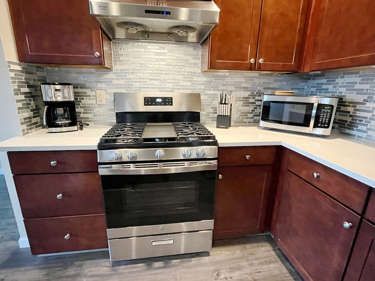 A modern kitchen is showcased with dark wood cabinets and sleek stainless steel appliances. A gas stove with an overhead exhaust hood is prominently displayed, alongside a microwave and coffee maker. Countertops are finished in white, with a textured backsplash adding visual interest.