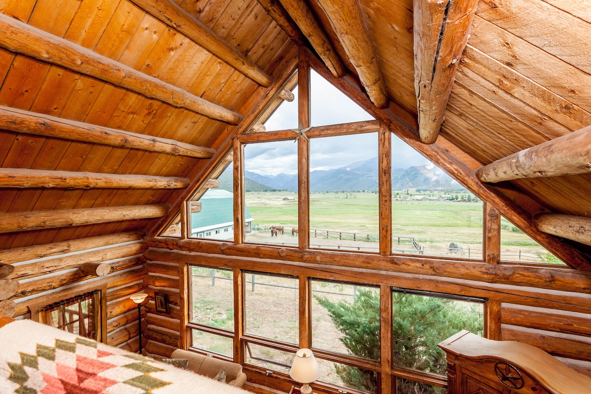 The interior of the log cabin features large windows that frame expansive views of the rolling green landscape and distant mountains. Natural wood beams accentuate the ceiling, and light streams in, creating a bright and inviting atmosphere.