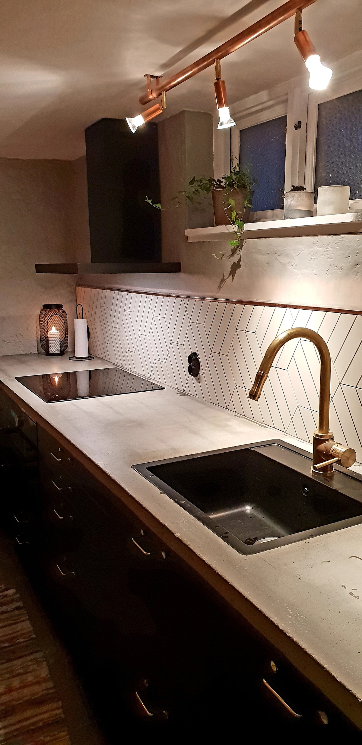 A modern kitchen features a sleek, minimalist design with a concrete countertop and dark cabinetry. Stylish fixtures in brass add a touch of elegance. A minimalist light fixture illuminates the space, highlighting geometric backsplash tiles. Natural light filters through a window beside a small plant.