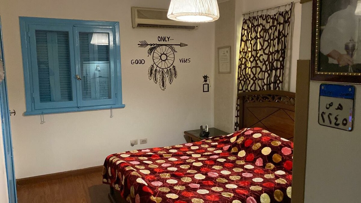 A spacious bedroom features a large bed covered with a colorful polka dot blanket. A dreamcatcher design is displayed on the wall, accompanied by blue shutters on a window. Soft lighting is provided by a pendant lamp, complementing the patterned curtains.