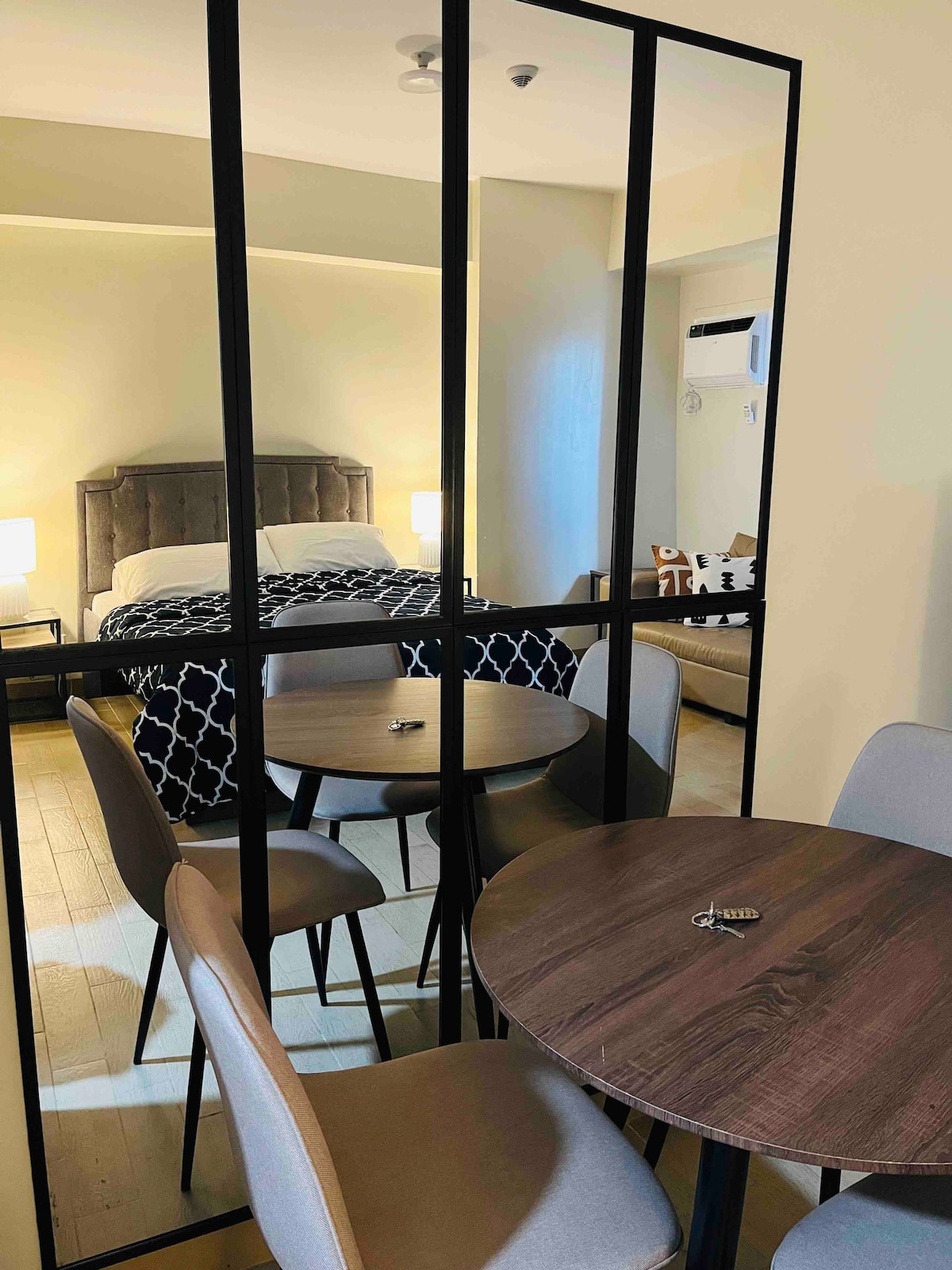 A modern dining area features a round wooden table surrounded by four upholstered chairs. A large mirror reflects a neatly made bed with a patterned blanket in the background, while soft lighting from bedside lamps enhances the ambiance.