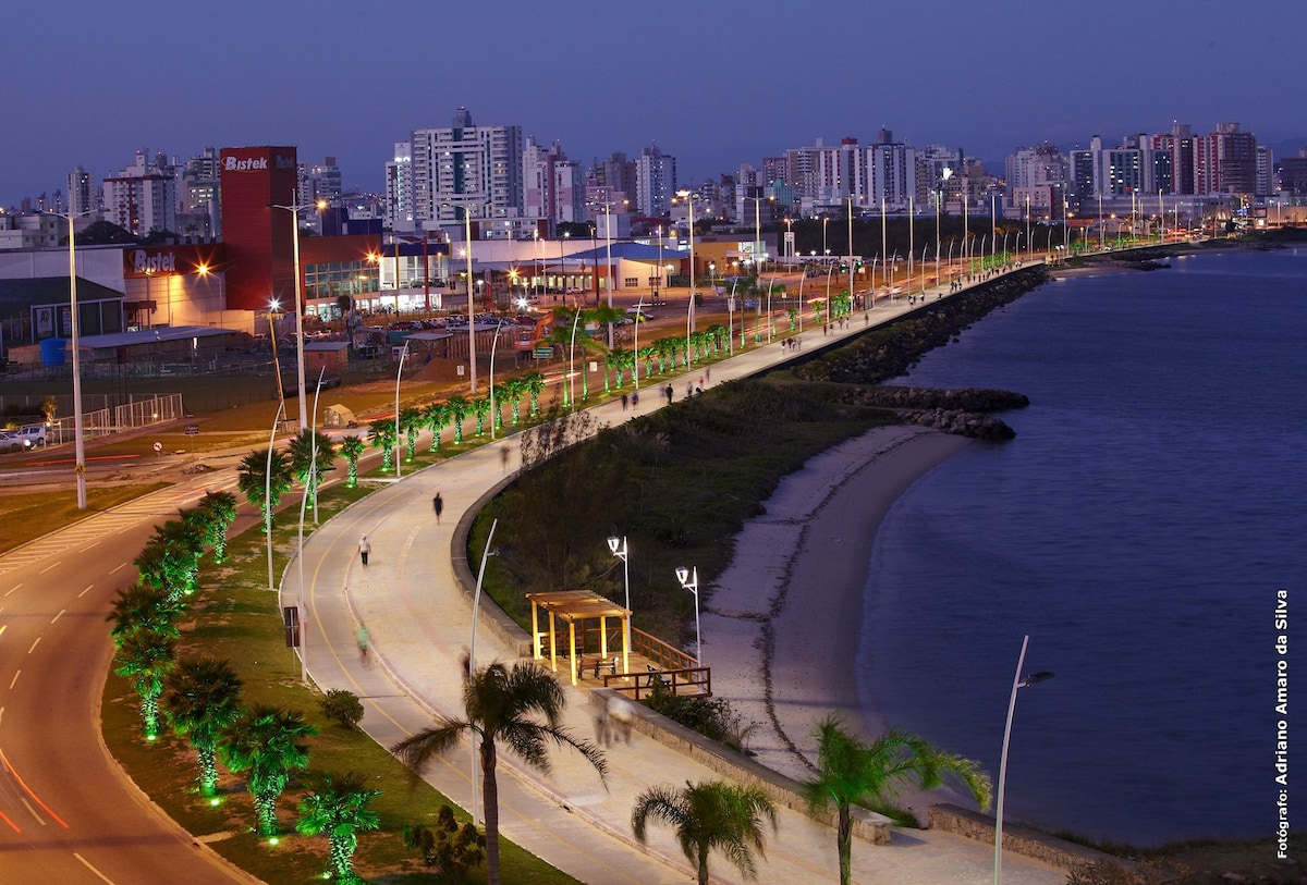 The image captures a coastal promenade illuminated by soft lights along the walkway. Lush landscaping features palm trees, while the beach curves gently beside the water. City buildings rise in the background against a twilight sky, contributing to a peaceful ambiance.