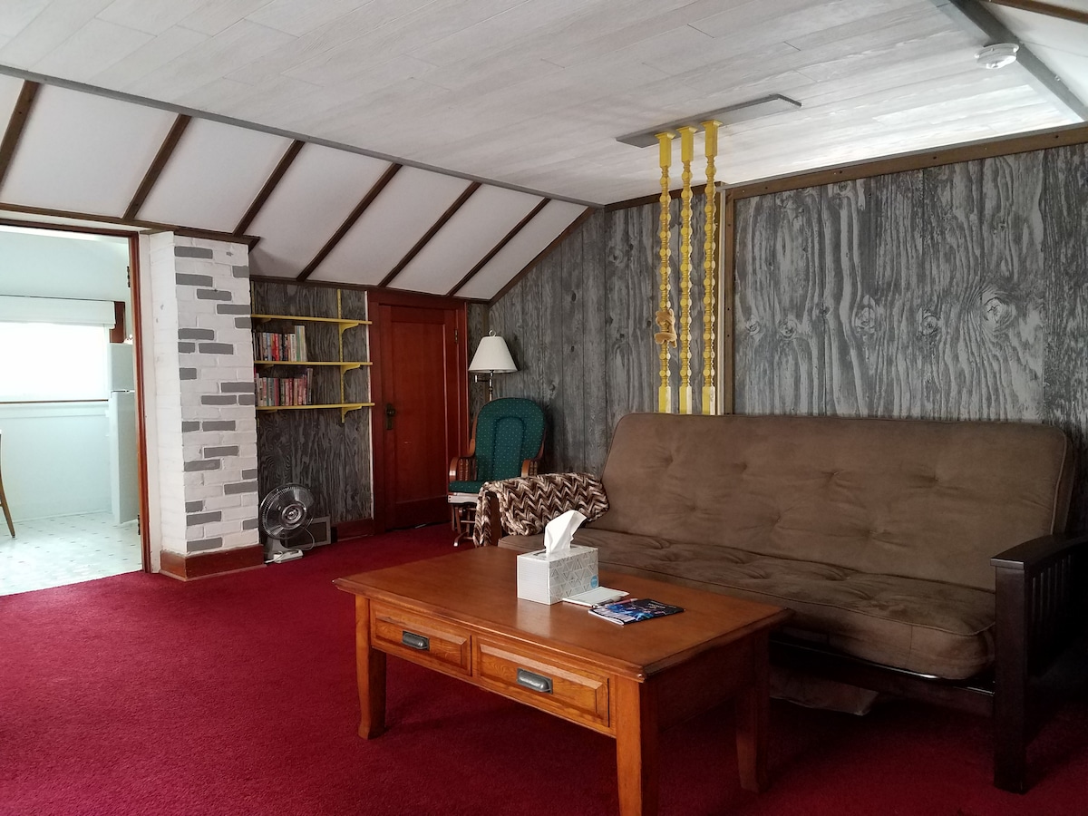 A cozy den is shown, featuring a futon couch, a wooden coffee table, and a fan. A bookshelf is positioned along the wall, and a green chair adds color to the space. The room is illuminated by natural light from the open kitchen visible in the background.