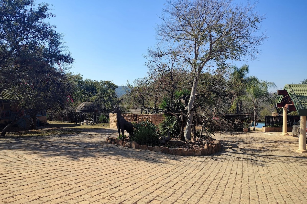 A spacious outdoor area features a paved surface surrounded by trees and shrubs, with a stone feature in the center. A shaded seating area is visible in the background, alongside a structure that appears welcoming and harmonious with the surrounding landscape.