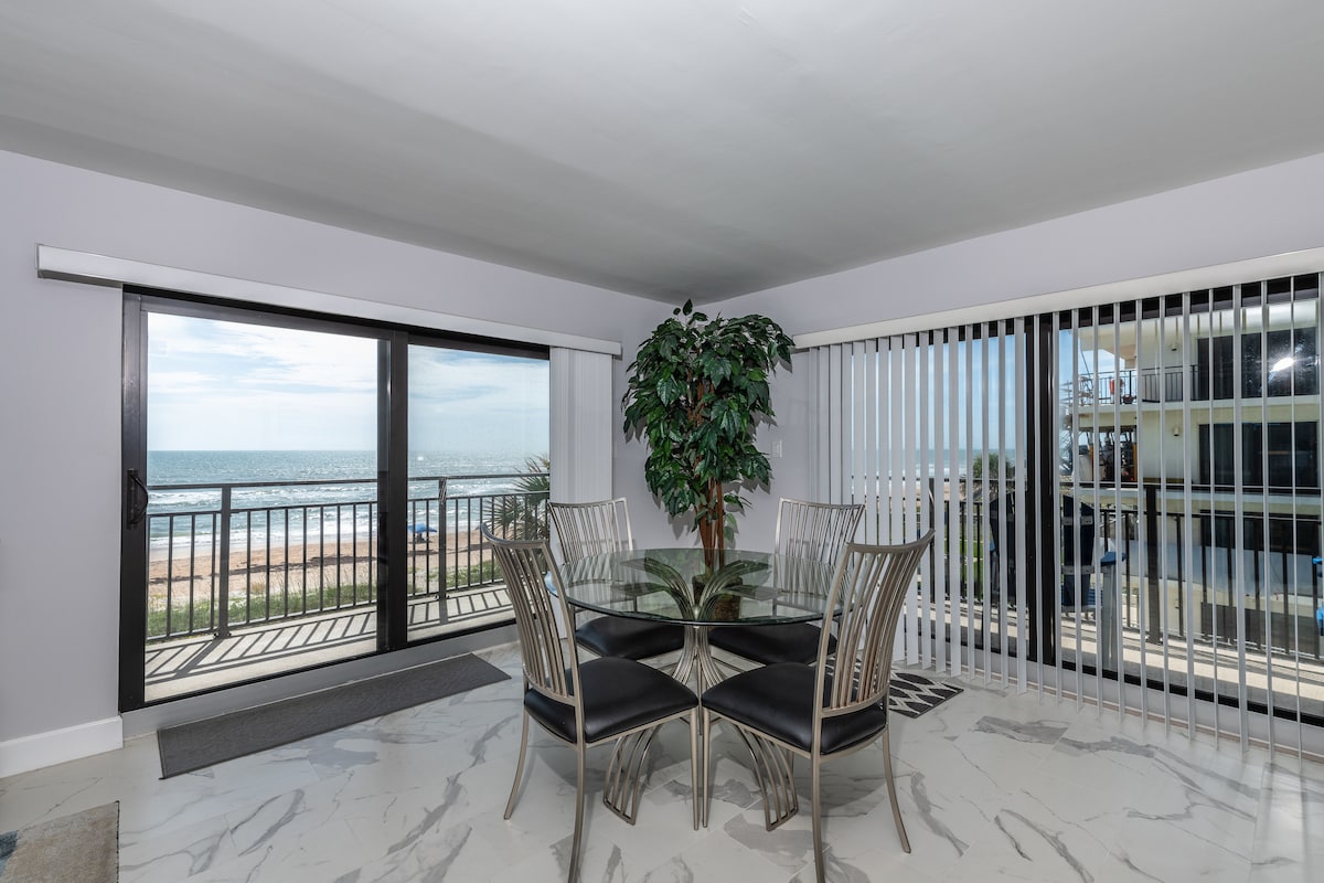 A dining area features a glass table surrounded by six metal chairs. Large sliding glass doors provide expansive views of the beach, while vertical blinds allow for control of natural light. A decorative plant adds a touch of greenery to the space.