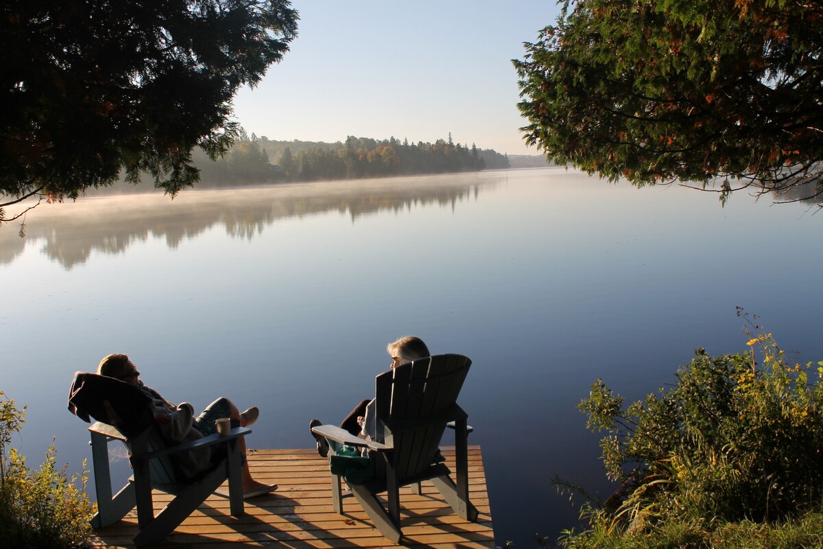 Two individuals relax on a private dock, seated in Muskoka chairs and facing a serene lake. The calm water reflects the morning light, with lush greenery framing the scene. Mist subtly hovers above the surface, adding to the tranquil atmosphere.