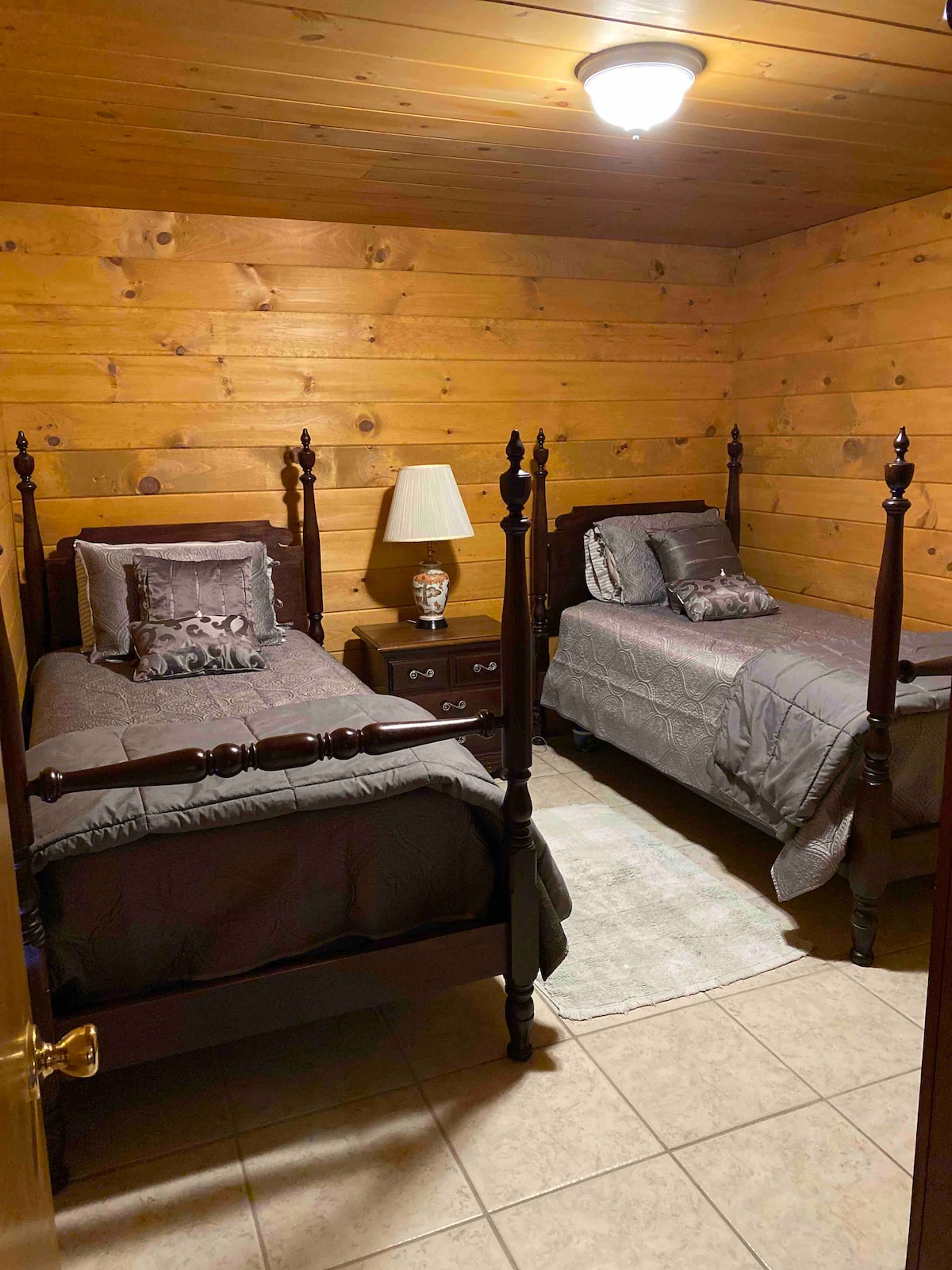 The room features two wooden beds with decorative bed linens and pillows. A bedside table with a lamp is positioned between the beds. The walls are finished with wood paneling, and soft lighting is provided by a ceiling fixture.