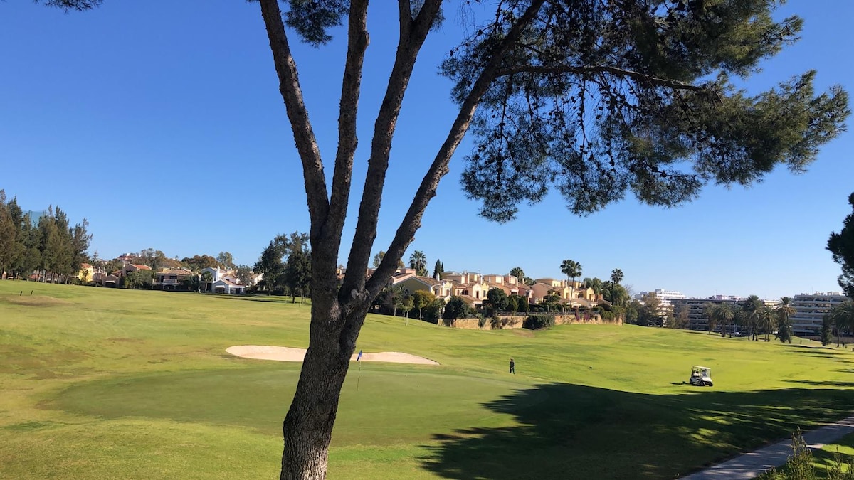 A serene golf course landscape is depicted, featuring lush green grass and a sand bunker to the left. Trees frame the scene, with residential buildings visible in the background. A clear blue sky complements the tranquil outdoor setting.