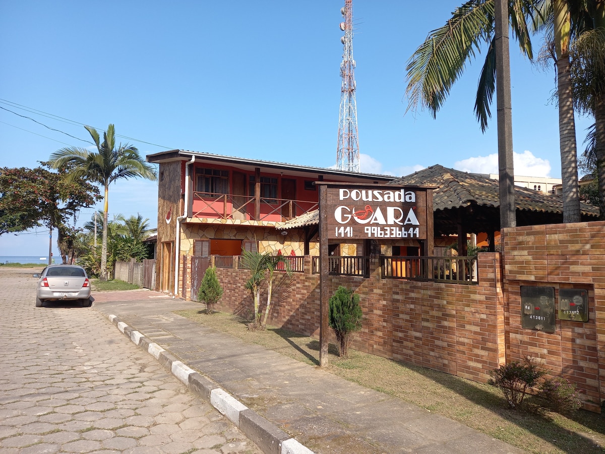 The entrance to the Pousada is characterized by a brown brick wall, complemented by green palm trees. A wooden sign displaying 'Pousada Guará' hangs prominently. The facade of the building features a mix of warm tones, with a rustic roof and clear blue skies in the background.