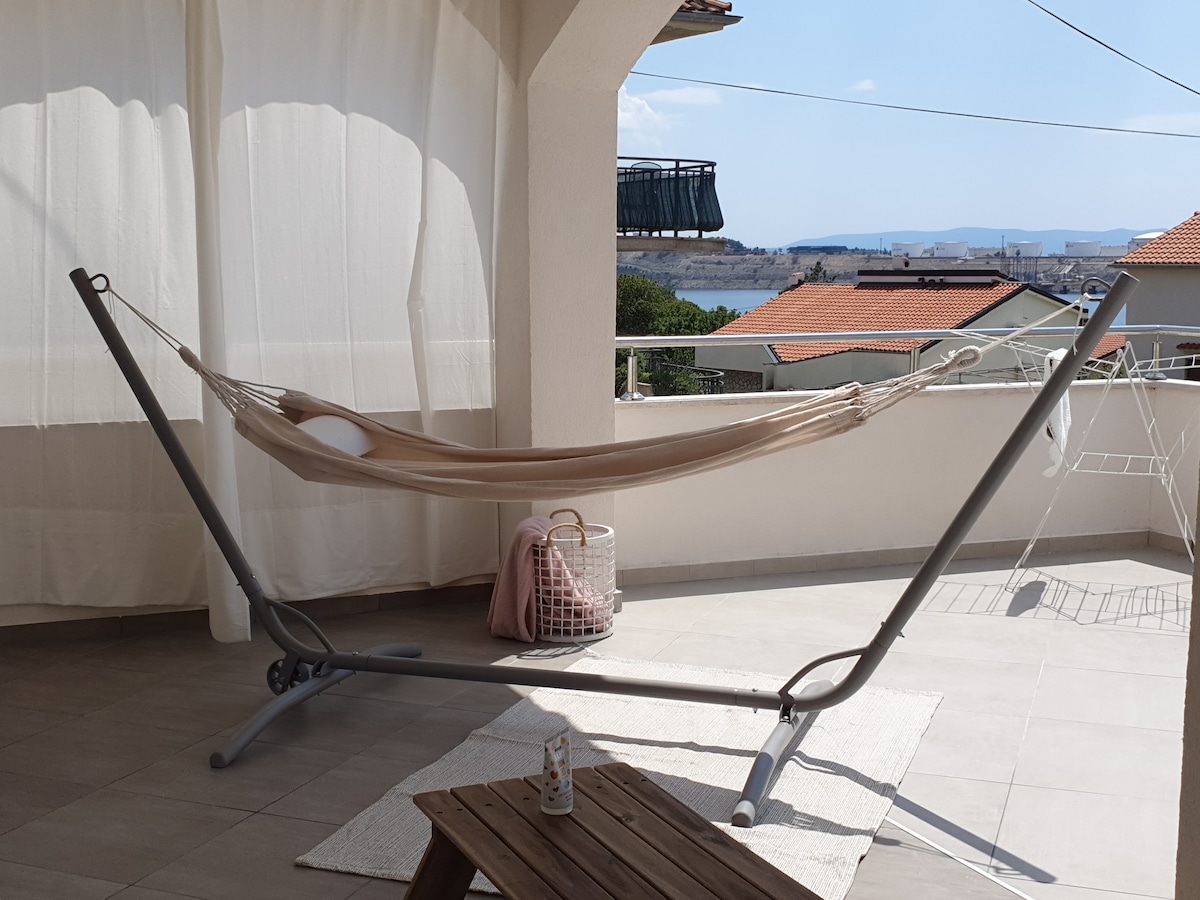A spacious terrace features a light-colored hammock suspended from a gray stand, accompanied by a small wooden table. A soft area rug is laid on the tiled floor, while a white basket and a decorative item are placed nearby. The sea view is visible in the background.