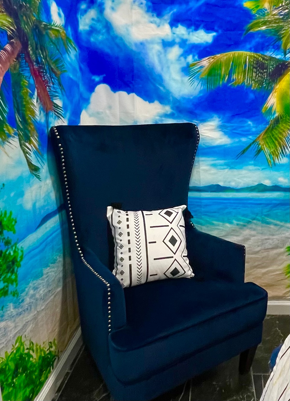 A tall, navy blue armchair is positioned against a vibrant mural depicting a tropical beach scene with palm trees and a bright sky. A patterned black and white pillow adds a contrasting element to the chair.