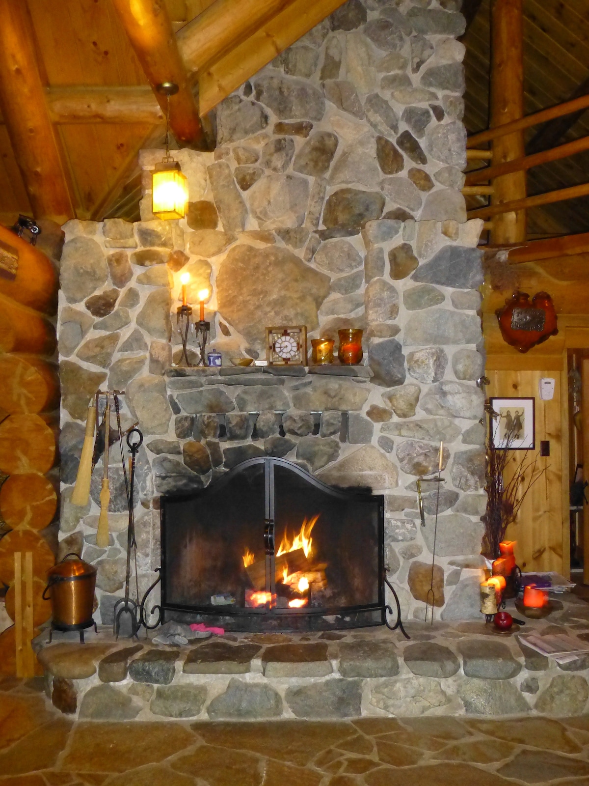 A large stone fireplace commands attention in the main living area, featuring a rustic design with a black metal screen. Flames flicker in the hearth, surrounded by various decorative items and candles, enhancing the cozy ambiance of the log home.