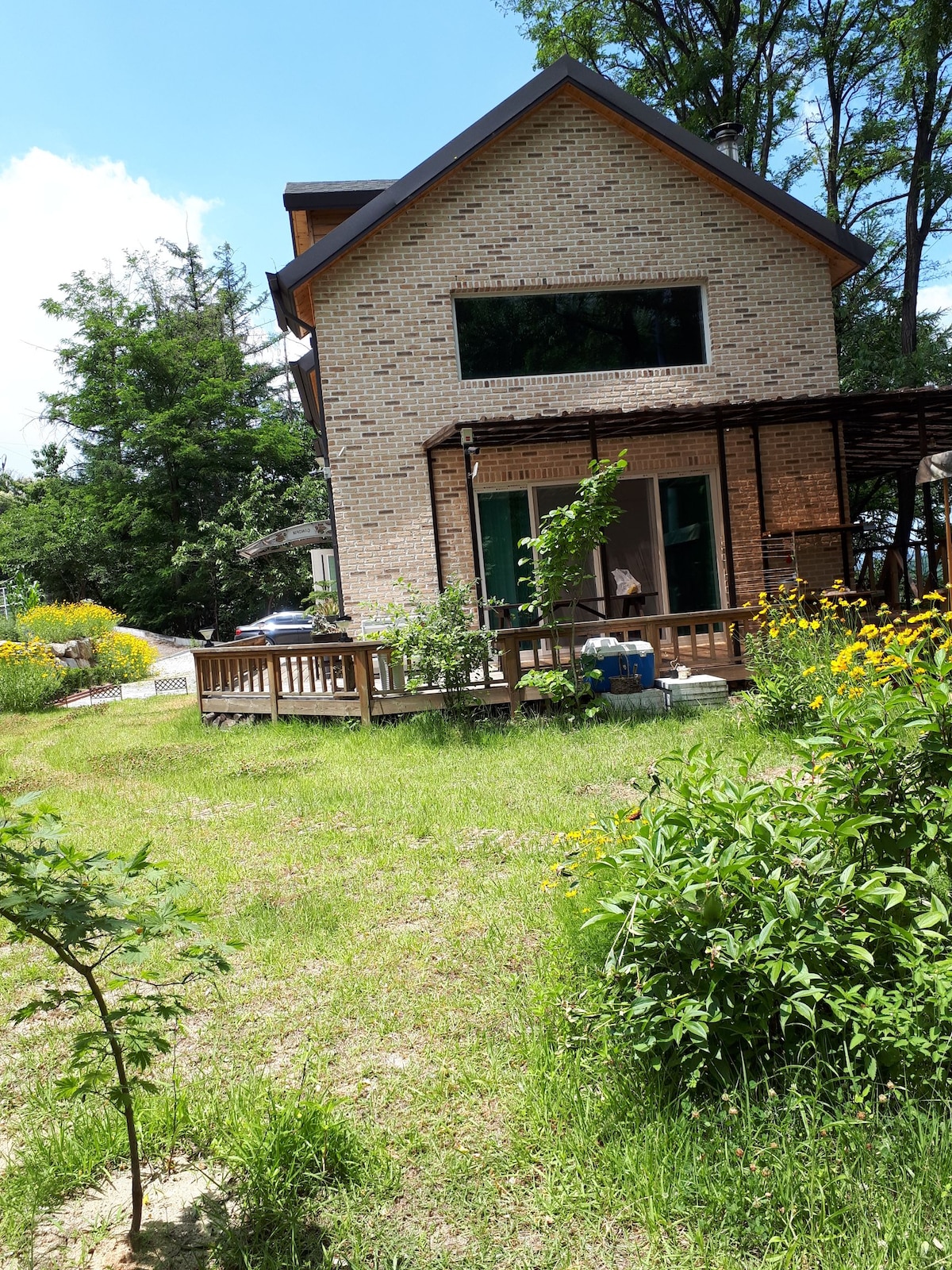 A two-story house is surrounded by a well-maintained garden, featuring vibrant flowers and greenery. A wooden deck is visible, providing an outdoor seating area. Large windows allow natural light to fill the interior, while mature trees offer shade nearby.