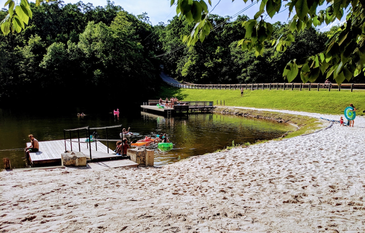A sandy beach area leads to a calm community lake surrounded by lush trees. Docks are visible, with people relaxing and kayaking on the water. Greenery stretches along the shoreline, enhancing the serene environment.