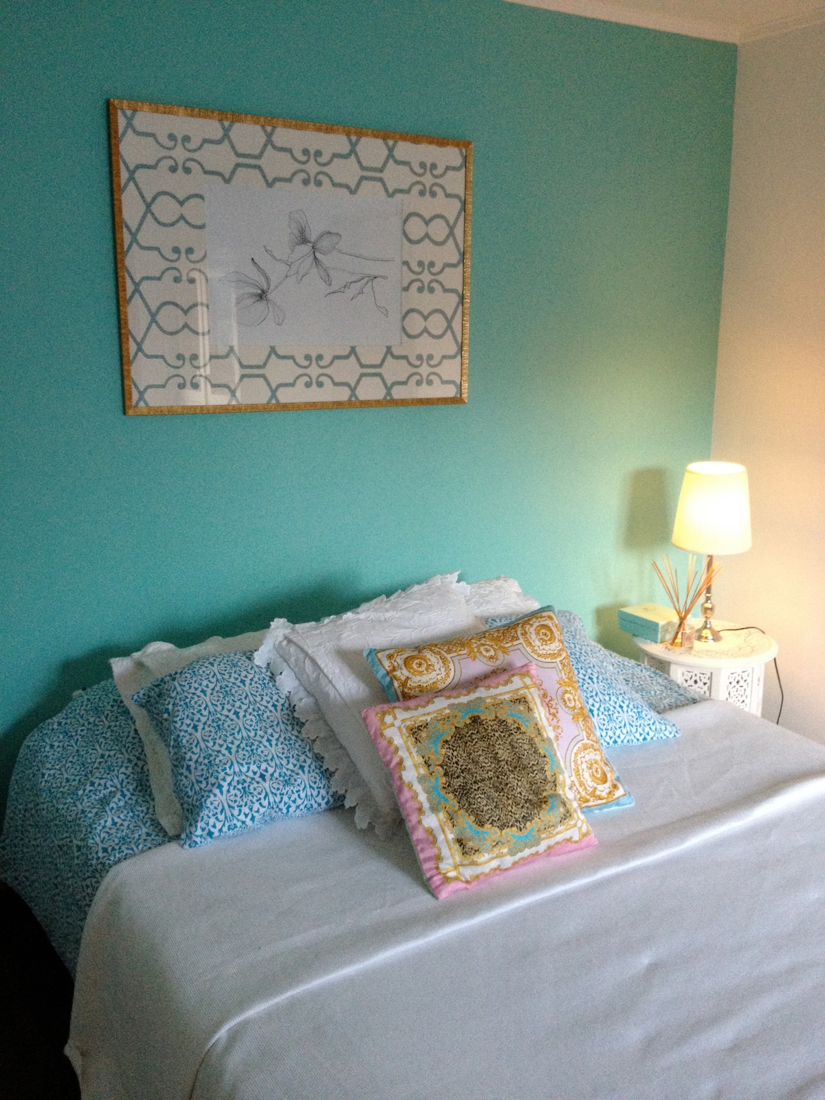 A cozy sleeping area features a spacious bed adorned with decorative pillows in various patterns. A soothing aqua wall provides a calming backdrop, while a bedside table with a lamp adds gentle illumination, creating a serene ambiance for relaxation.
