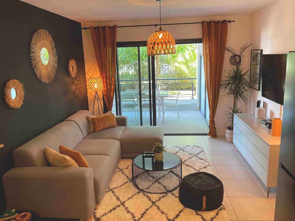A modern living area features a light grey sofa and round coffee table on a textured rug. Natural light is filtered through large sliding doors, leading to an outdoor terrace. Decorative elements include wall art and a potted plant, enhancing the relaxed ambiance.