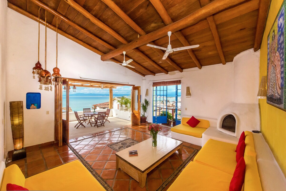 A spacious living area features a bright yellow sofa and a coffee table, with large windows that frame a sea view. Light wood beams span the ceiling, creating a warm ambiance. A small dining area with a table and chairs is visible through the open doors.