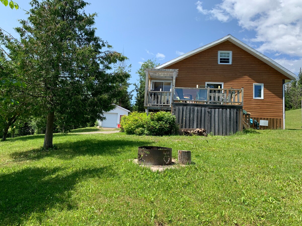 A rustic chalet is shown nestled in a green landscape, featuring a wooden deck with seating and a fire pit in the foreground. The structure is surrounded by trees and a spacious yard, with a garage visible in the distance.