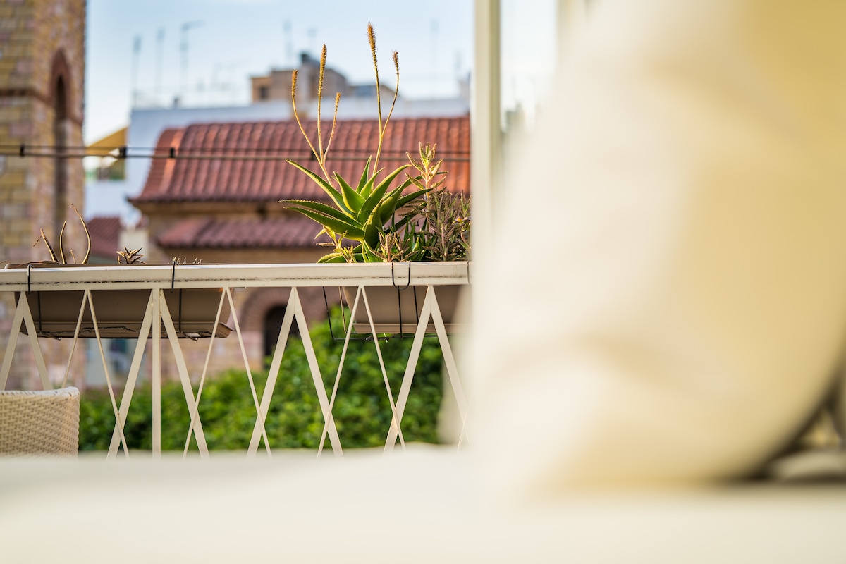 A balcony view is presented with a focus on a lush green plant juxtaposed against a backdrop of terracotta rooftops. Soft furnishings are visible in the foreground, creating a sense of comfort and tranquility in this outdoor space.