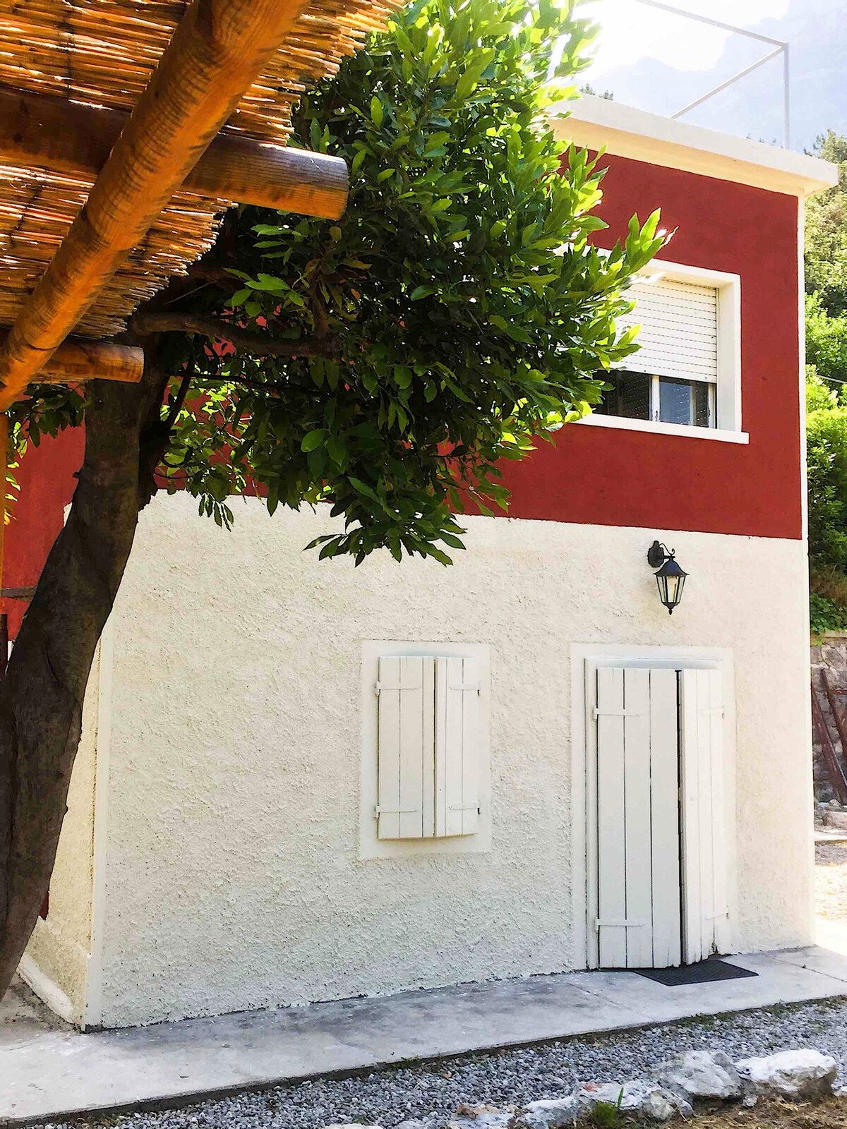 The exterior of Casa Cantoniera features a two-story building with a striking red facade and light-colored textured walls. White shutters adorn the windows, while a lantern hangs beside the door, complemented by a tree providing shade at the entrance.