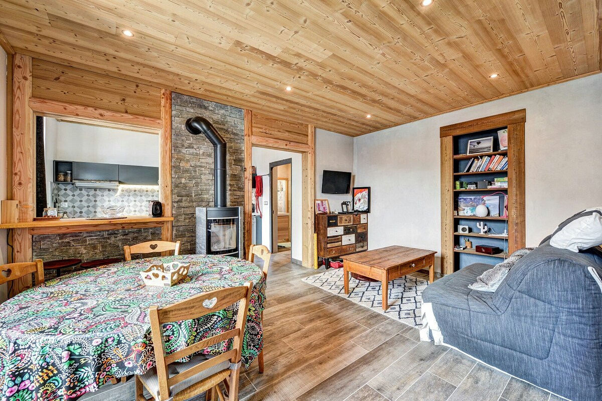 The open living space features warm wooden paneling and a cozy seating area. A round table with a colorful tablecloth occupies the center, while a comfortable sofa is positioned nearby. A decorative rug anchors the space, and a stone fireplace adds character to the room.