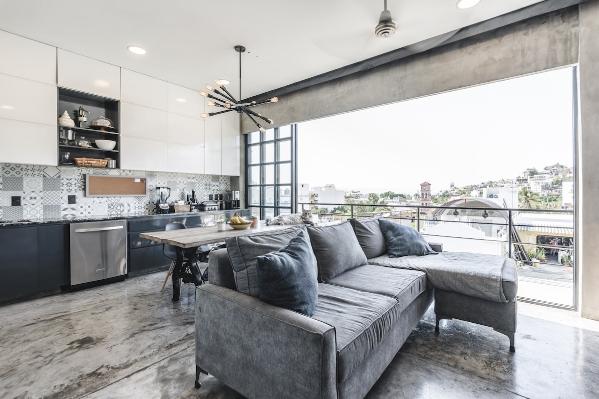 A modern kitchen space features sleek cabinetry and a large island, complemented by stylish light fixtures. A spacious sectional sofa is positioned near large glass doors, offering expansive views of the surrounding area. Natural light fills the room, enhancing the open layout.