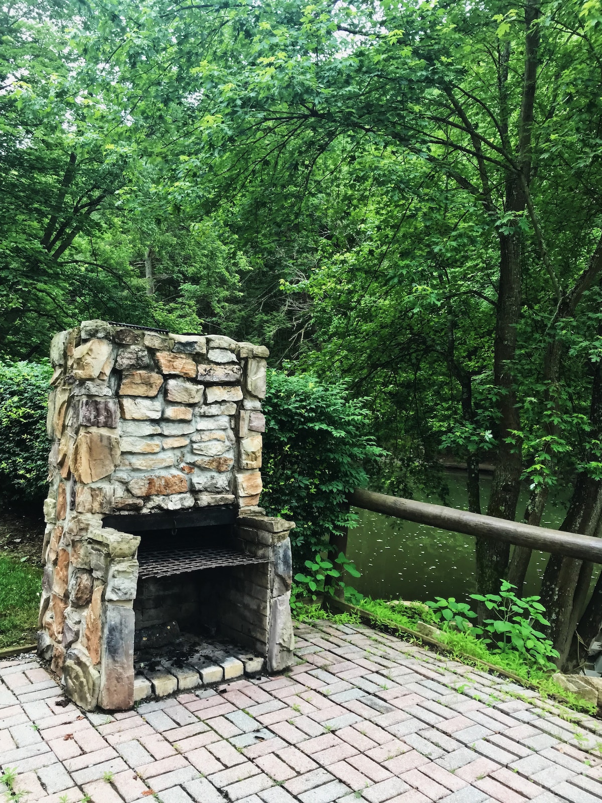 A stone fireplace is situated on a paved patio, surrounded by lush greenery and trees. The tranquil river is visible in the background, enhancing the outdoor atmosphere for gatherings and relaxation.