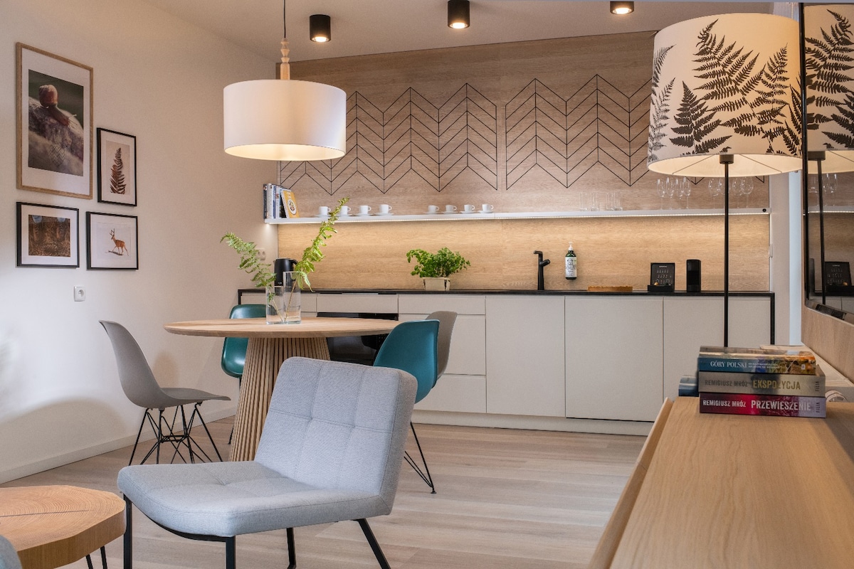 An open living space features a circular dining table surrounded by colorful chairs, complemented by a modern gray armchair. A wall showcases geometric patterns, while shelves display small plants and tableware. Two light fixtures provide illumination, enhancing the room's contemporary aesthetic.