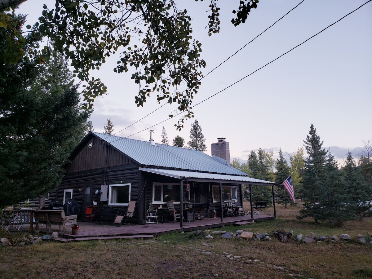 A rustic cabin sits surrounded by greenery and trees, featuring a covered porch with several seating areas. An American flag gently flutters in the breeze, complementing the peaceful outdoor setting. The sloped roof and chimney add character to this inviting retreat.