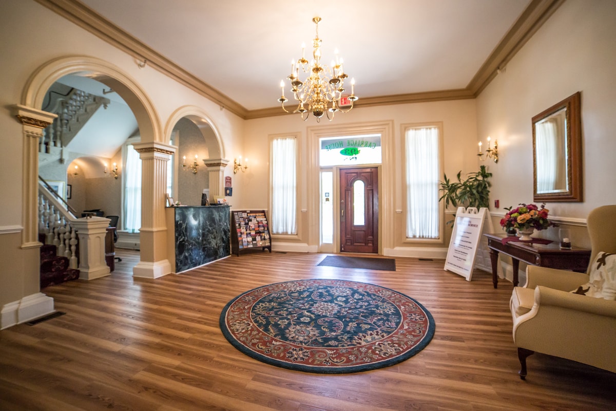 The welcoming lobby features polished wooden floors and a centrally placed decorative area rug. A chandelier hangs overhead, illuminating the space. Light streams through large windows, highlighting the elegant furnishings, including a chair and a side table adorned with flowers.