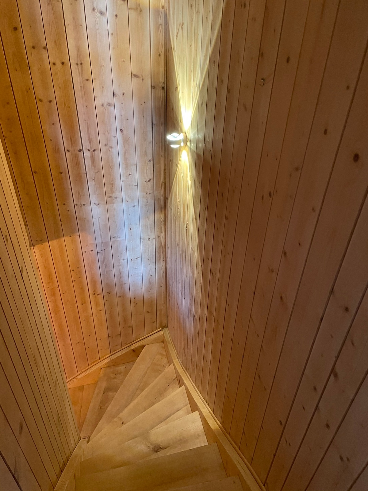 A wooden spiral staircase is seen, featuring warm wooden planks and walls lined with matching timber. Soft lighting illuminates the space, highlighting the texture of the wood. The curved design leads the eye downward, suggesting an inviting transition to the lower level.