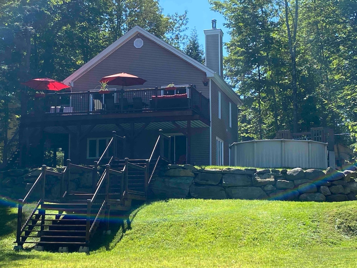 A spacious chalet is situated on a grassy slope, featuring a deck with red umbrellas. A staircase leads from the lawn to the entrance. A circular above-ground pool is positioned nearby, surrounded by trees, providing a serene outdoor setting.