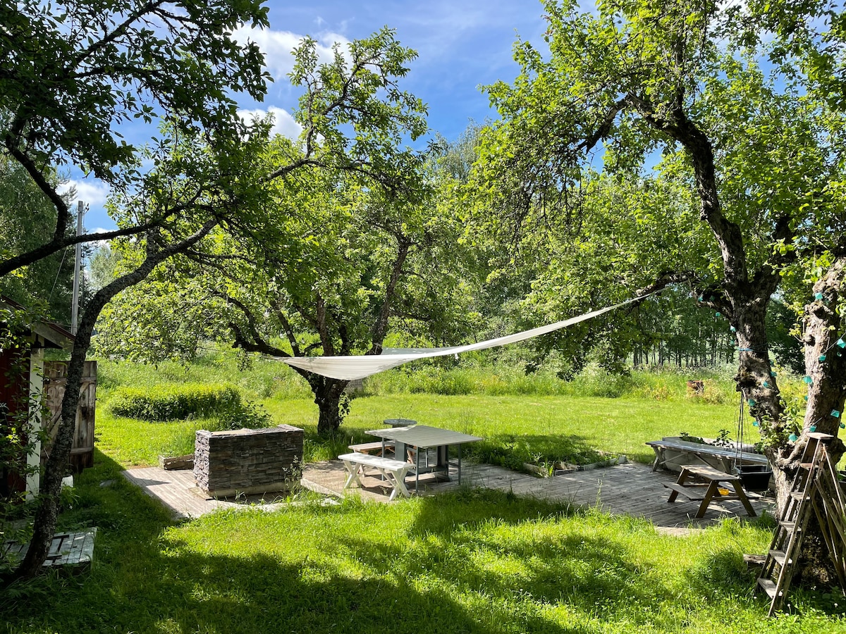 An outdoor area features a spacious picnic table beneath shade from trees, with a sail cloth stretched overhead. Grass surrounds the seating area, and a wooden deck is visible. Additional seating can be seen nearby, creating a welcoming outdoor space for relaxation.