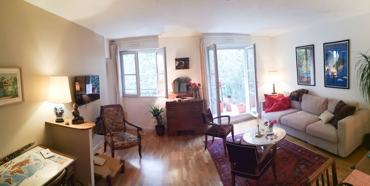 A spacious living area features a comfortable sofa, two armchairs, and a coffee table on a patterned rug. Natural light enters through double windows, overlooking greenery. A side table holds a vase of flowers, and an art piece adorns the wall, contributing to a relaxing setting.