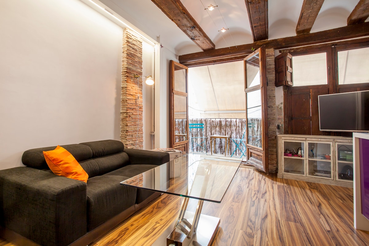A spacious living area features wooden beams and a large window that opens to a balcony. A comfortable dark sofa with an orange cushion sits in front of a glass coffee table. The warm wood flooring complements the rustic brick accent wall.