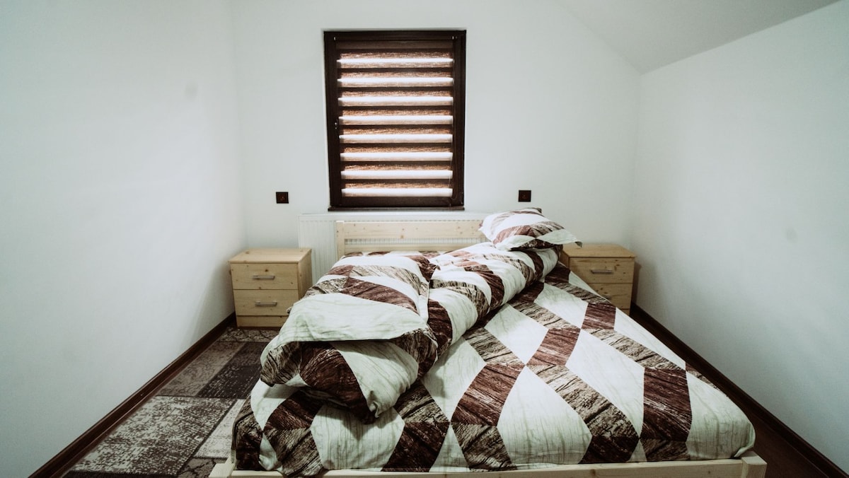 A cozy bedroom features a spacious bed dressed in a patterned quilt and multiple pillows. Two wooden nightstands are positioned on either side of the bed. Natural light filters through a window with layered blinds, illuminating the light walls and flooring.