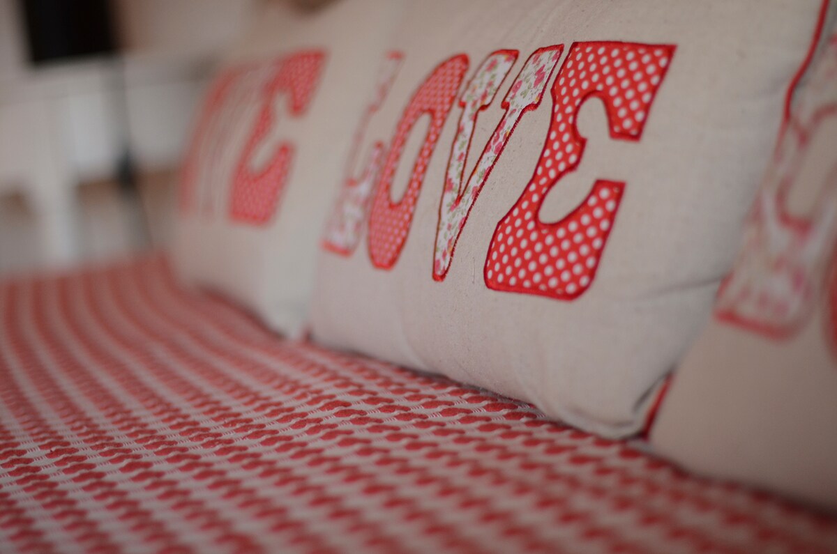 Soft pillows are arranged on a patterned bedspread featuring red and white checkered designs. The pillows, with bold letters spelling 'LOVE,' add a warm touch to the inviting sleeping area.