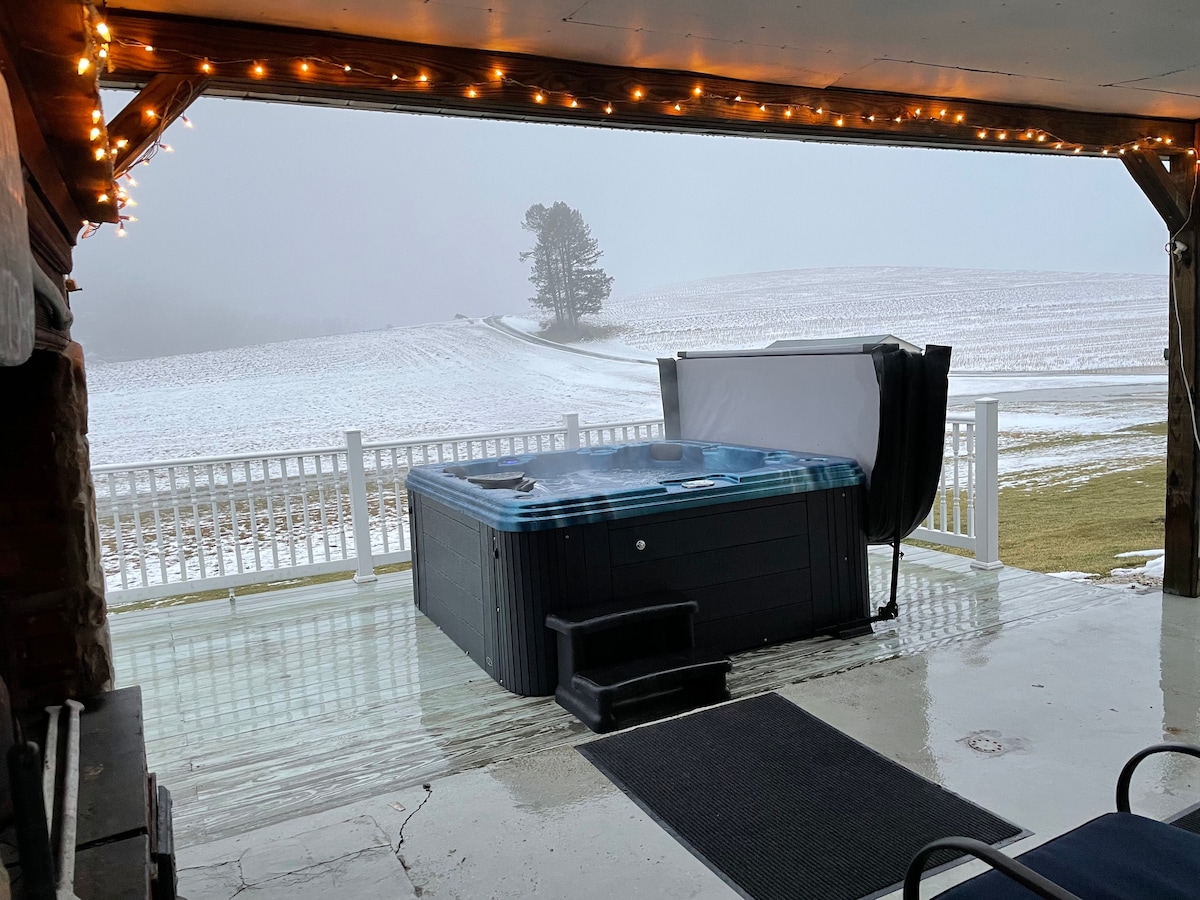 A covered deck area features a hot tub with steam rising gently above the water. String lights are visible overhead, creating a cozy ambiance. Outside, a winter landscape is seen, with rolling hills partially shrouded in fog and a snow-dusted path leading away.