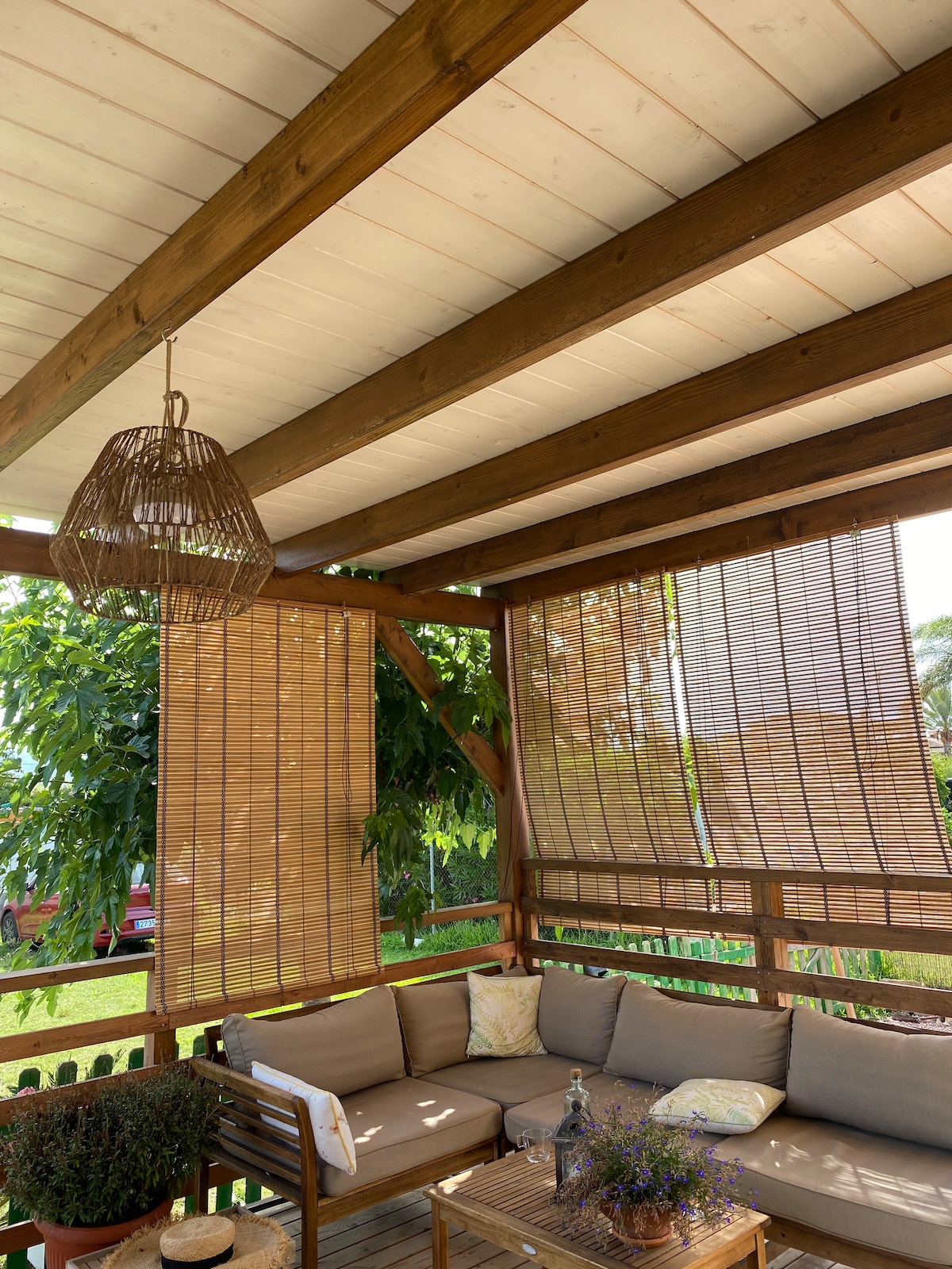 A spacious porch is showcased with natural wooden beams overhead and woven curtains for privacy. Comfortable seating is arranged around a wooden table, accented by a decorative vase with flowers. Soft lighting is provided by a woven pendant lamp hanging above the seating area.