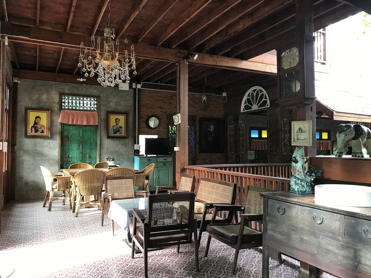 The interior space features traditional wooden beams and a variety of seating options, including wicker chairs and a dark wooden table. Decorative elements include wall art and vintage clocks, while large windows allow natural light to fill the room.