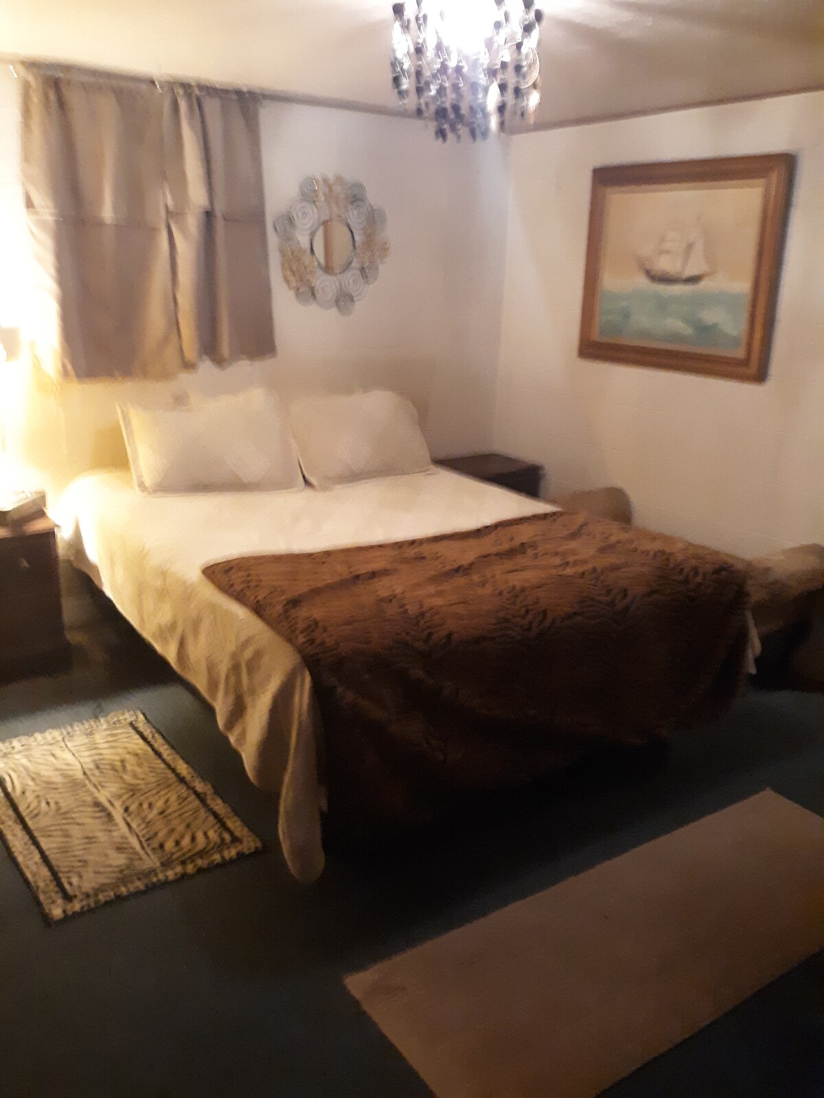 A cozy sleeping area features a spacious bed with a patterned brown blanket and two white pillows. Soft lighting is provided by a bedside lamp. A decorative mirror and a framed painting adorn the walls, adding character to the room.