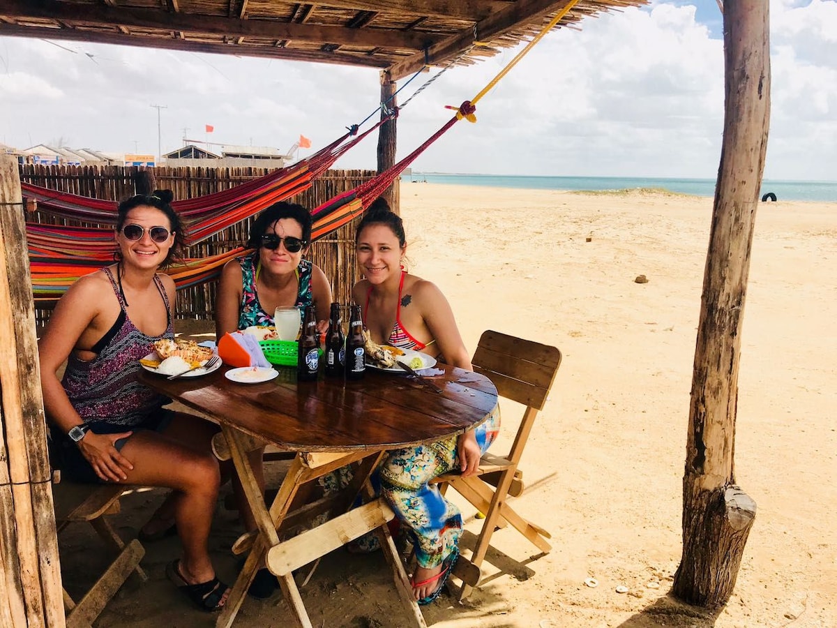 A shaded enramada at the beach shows three individuals seated at a wooden table, enjoying meals with beverages. Colorful hammocks are hung nearby, and the sandy beach extends toward the ocean, under a partly cloudy sky.